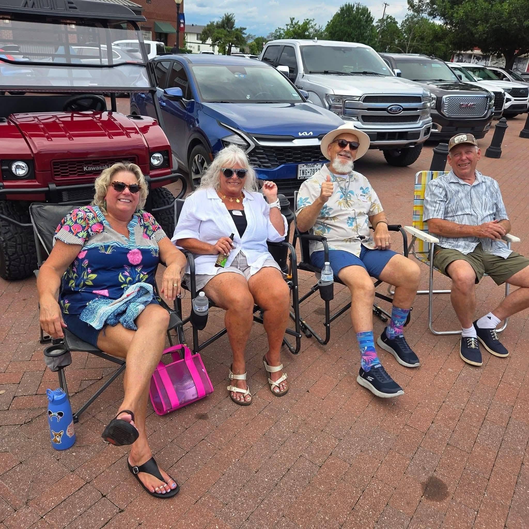 Four people seated in camp chairs on a brick street, with a golf cart and trucks behind them.