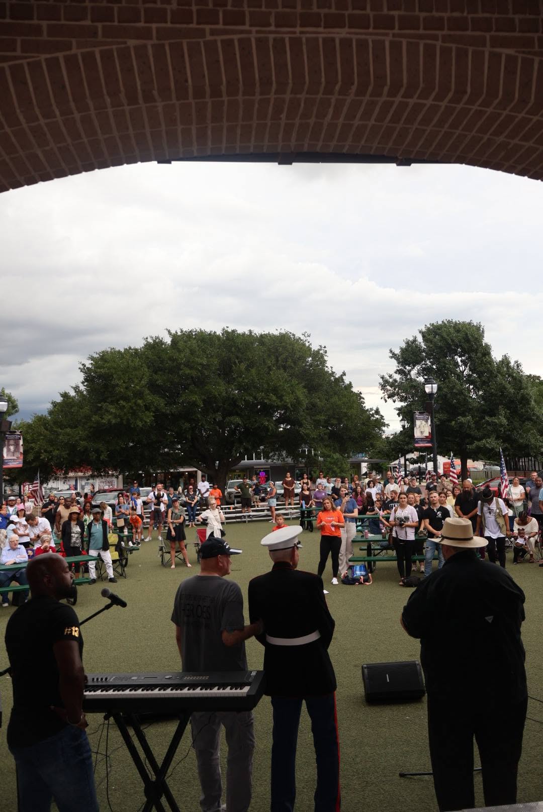 A crowd gathered on a lawn under trees, seen from the stage through a brick arch.