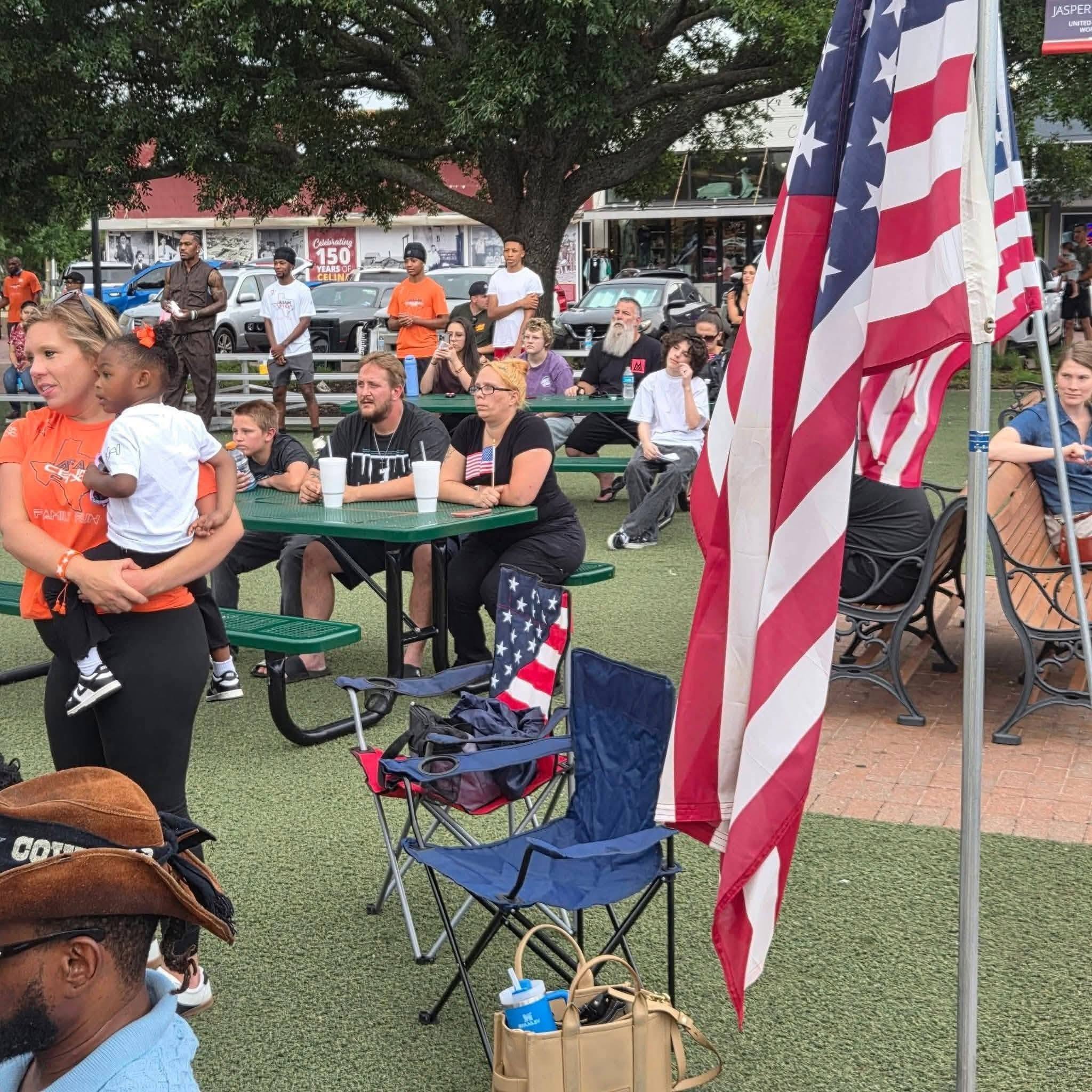 People seated at picnic tables under a tree, with an American flag in the foreground.