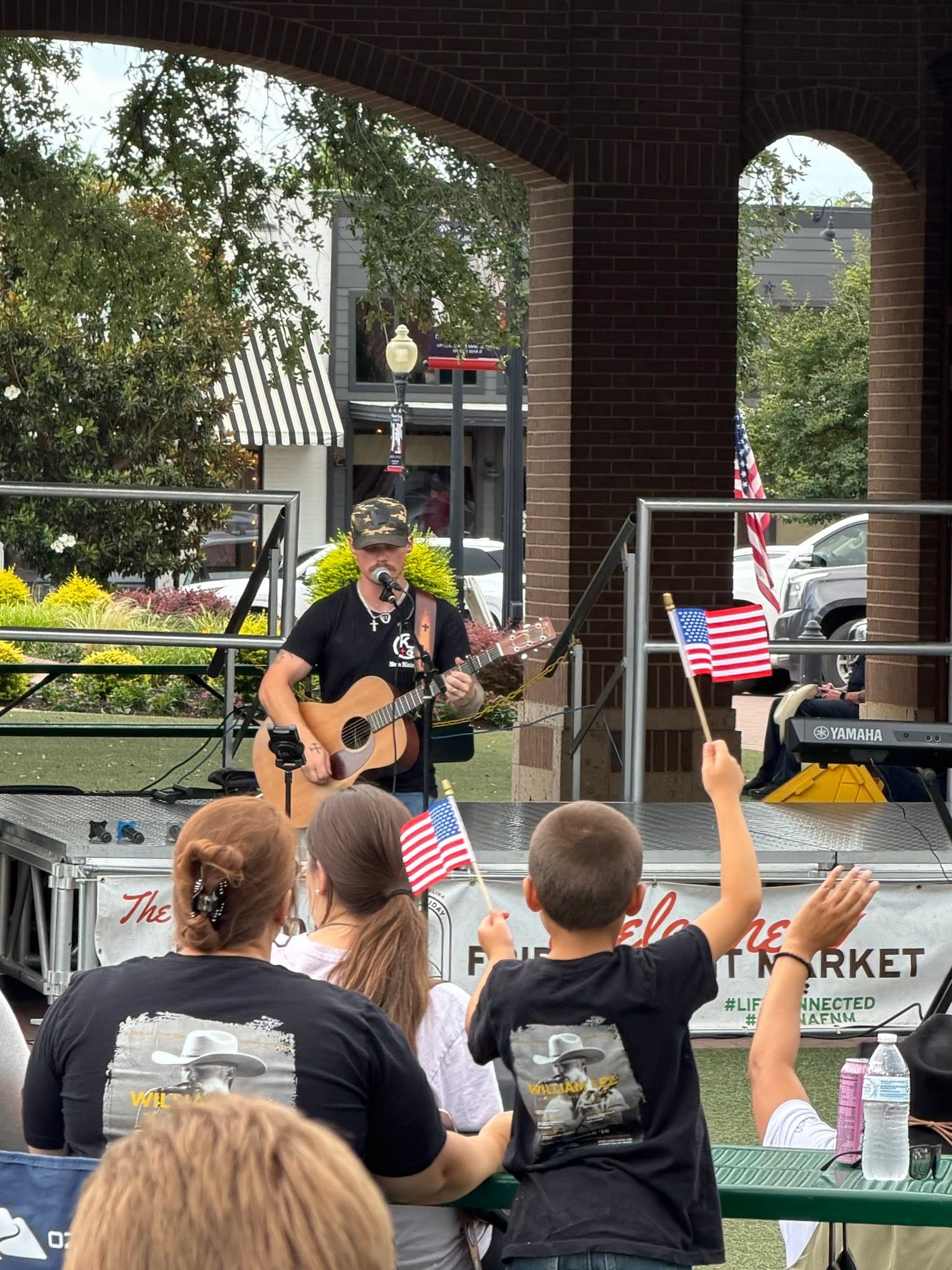 A man playing guitar under a pavilion while children hold small American flags.