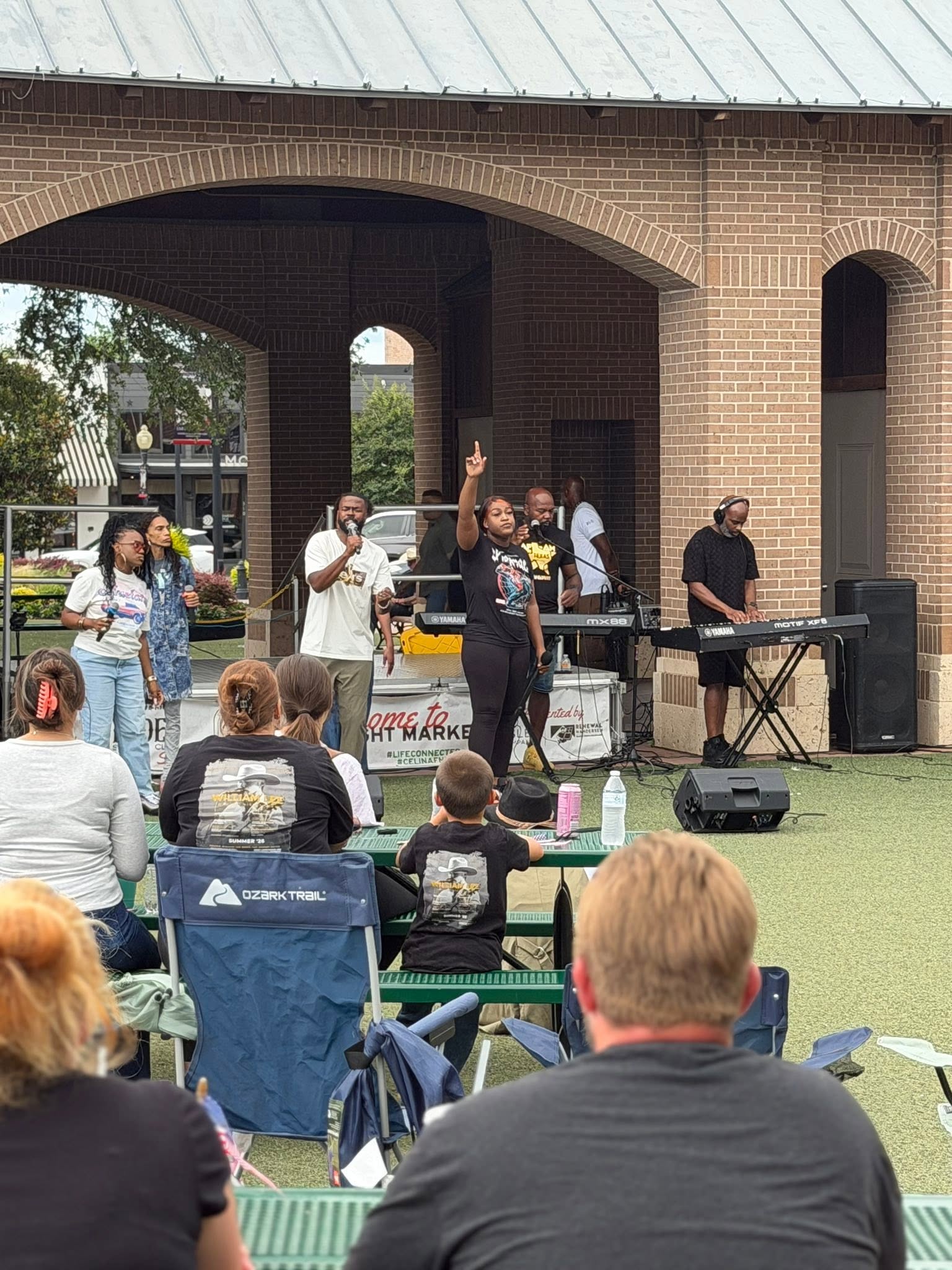 A woman singing under a brick pavilion, with a keyboard player and people seated at picnic tables.