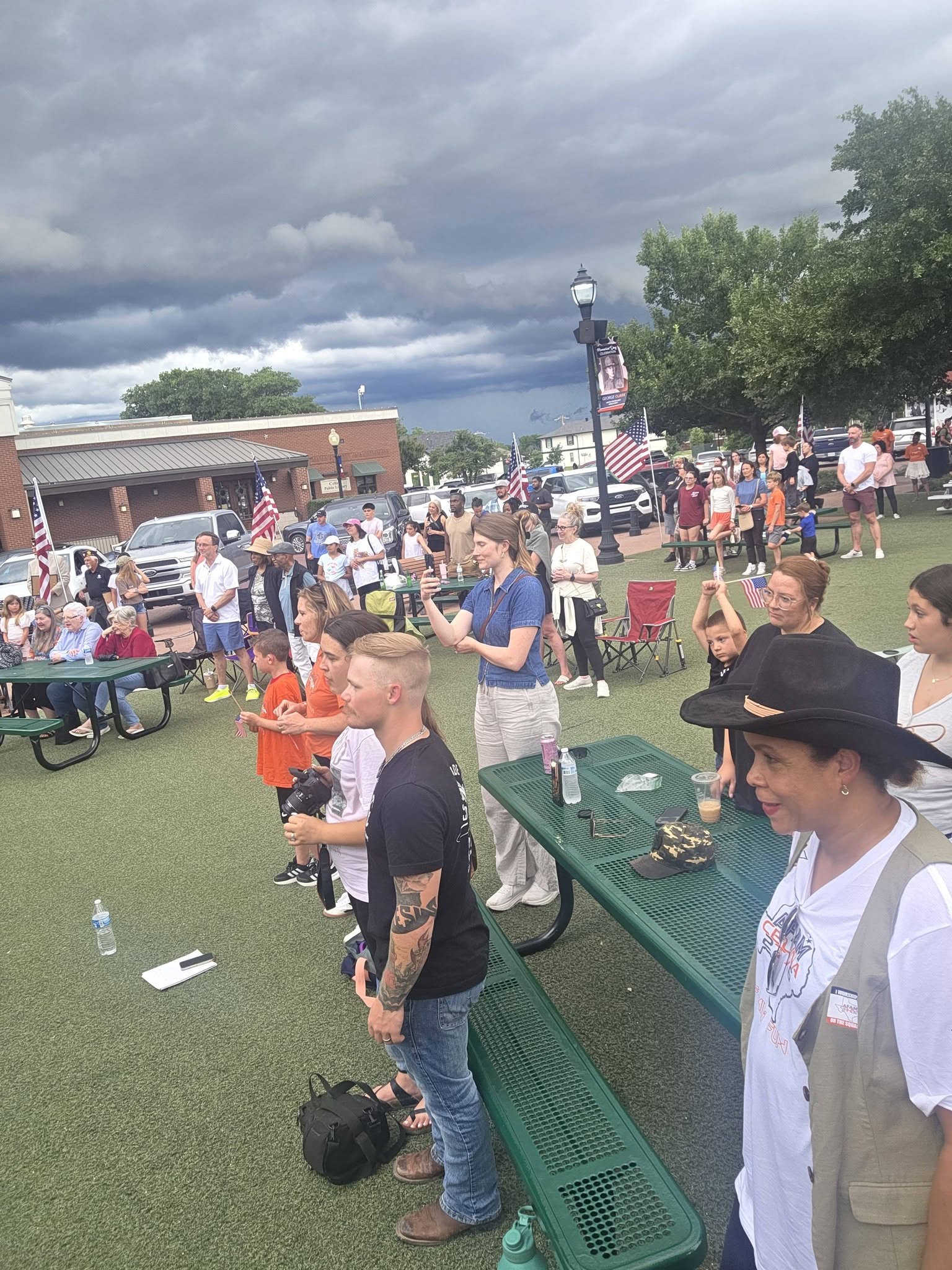 People standing and seated around picnic tables at an outdoor gathering, with flags behind them.