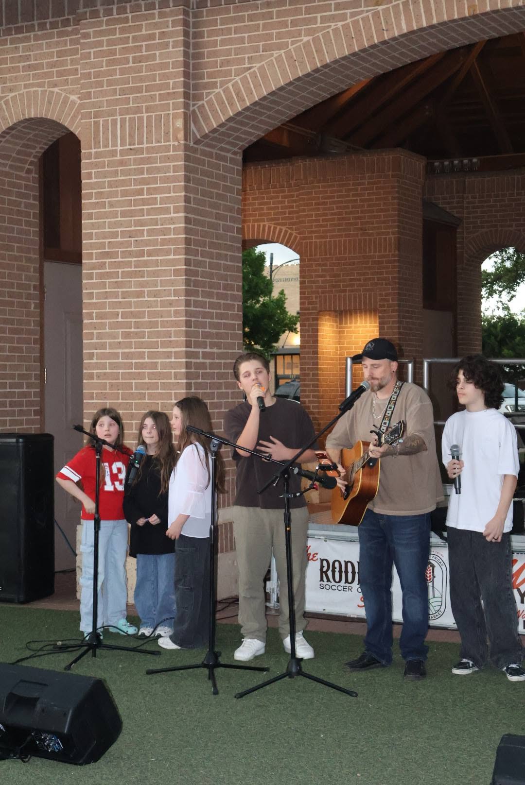 Young people at microphones under a brick pavilion, with a man playing guitar.